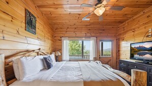 Un dormitorio de cabaña de madera con una cama, un ventilador de techo, un televisor y una ventana con vistas a árboles.