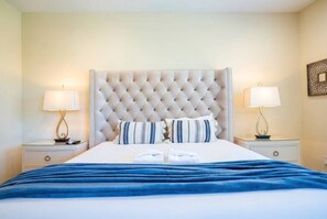 Een hotelkamer met een gestoffeerd hoofdeinde, twee nachtkastjes met lampen, een bed met witte lakens en een blauwe plaid.