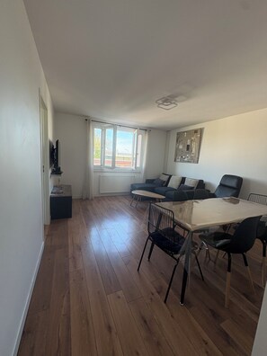A spacious living area with a sofa set, a dining table with chairs, a radiator, and a window with curtains.