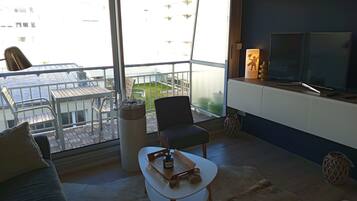 Een moderne woonkamer met een bank, een kleine tafel, een stoel, een televisie en een balkon met uitzicht op gebouwen.