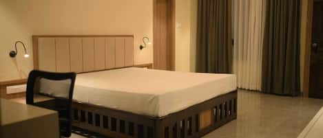 A hotel room with a bed, a desk, a chair, a nightstand, a lamp, a door, and a window with curtains.