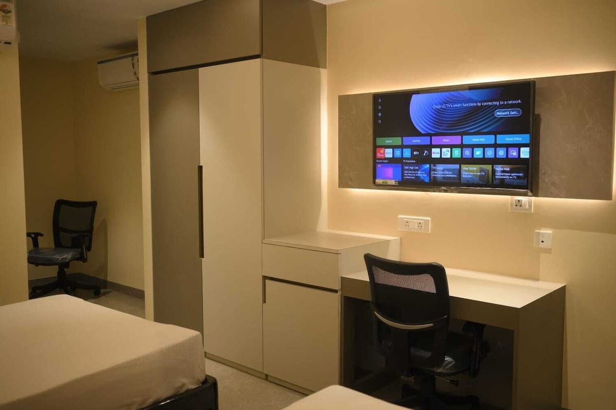 A hotel room with a desk, chair, and a TV mounted on the wall displaying various apps.