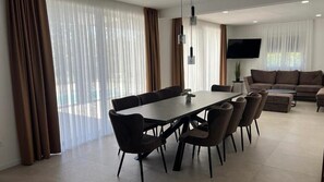 Una sala de estar moderna con comedor, ventanas amplias con cortinas, televisor de pantalla plana y un sofá.