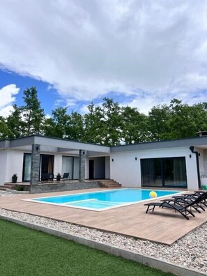 Una casa moderna con piscina, un patio con sillones de descanso y un césped bien cuidado.