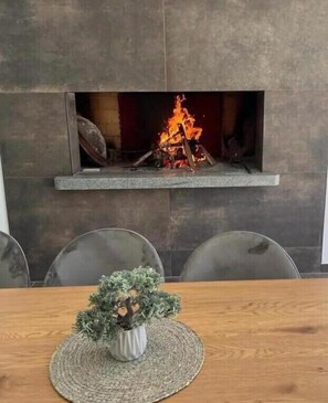 A fireplace with a fire, a dining table with chairs, and a small potted plant on the table.
