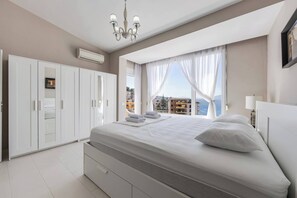 A bedroom with a large bed, white wardrobe, and a view of buildings and the sea. - Serenity by the Sea Duplex | Bougainville Resort by PikHost (Sarandë)