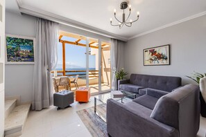 Een moderne woonkamer met een grijze bank, een glazen salontafel en uitzicht op de zee door een schuifpui.