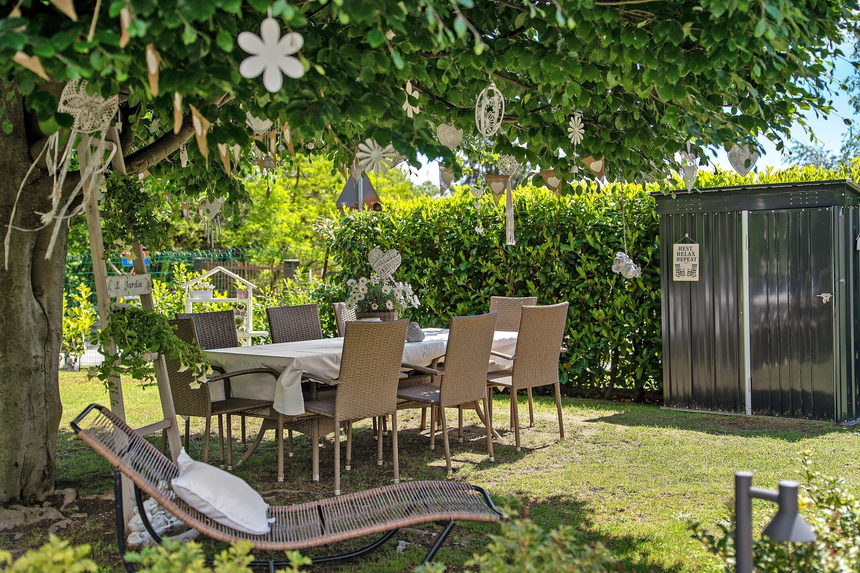 A garden setting with a table and chairs under a tree, a small shed, and a sign indicating a rest area.