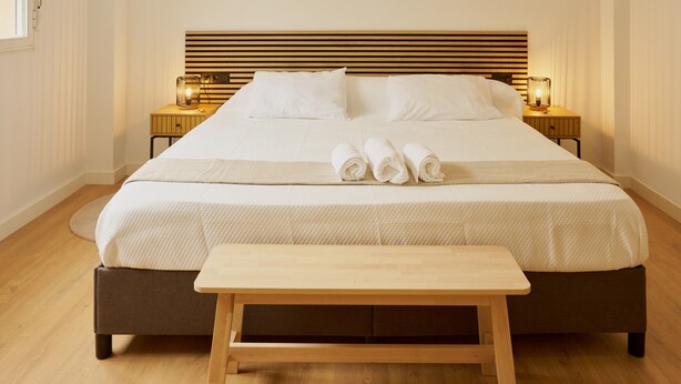 Cama bem arrumada com lençóis brancos, cabeceira de madeira, luminárias de cabeceira e um banco de madeira em um quarto de hotel.