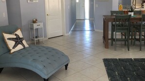 A living room with a blue sofa, a small white side table, a kitchen area with a dining table and chairs, and a hallway leading to another room. - Walk to Beach! Private Heated 3 bedroom Pool Home (Naples)