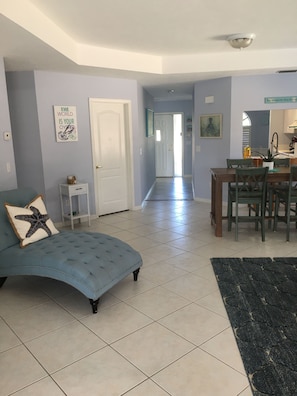 A living room with a blue sofa, a small white side table, a kitchen area with a dining table and chairs, and a hallway leading to another room. - Walk to Beach! Private Heated 3 bedroom Pool Home (Naples)