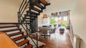 Un interior moderno con una escalera de madera, un comedor con sillas y una mesa, y vistas a través de grandes ventanales.