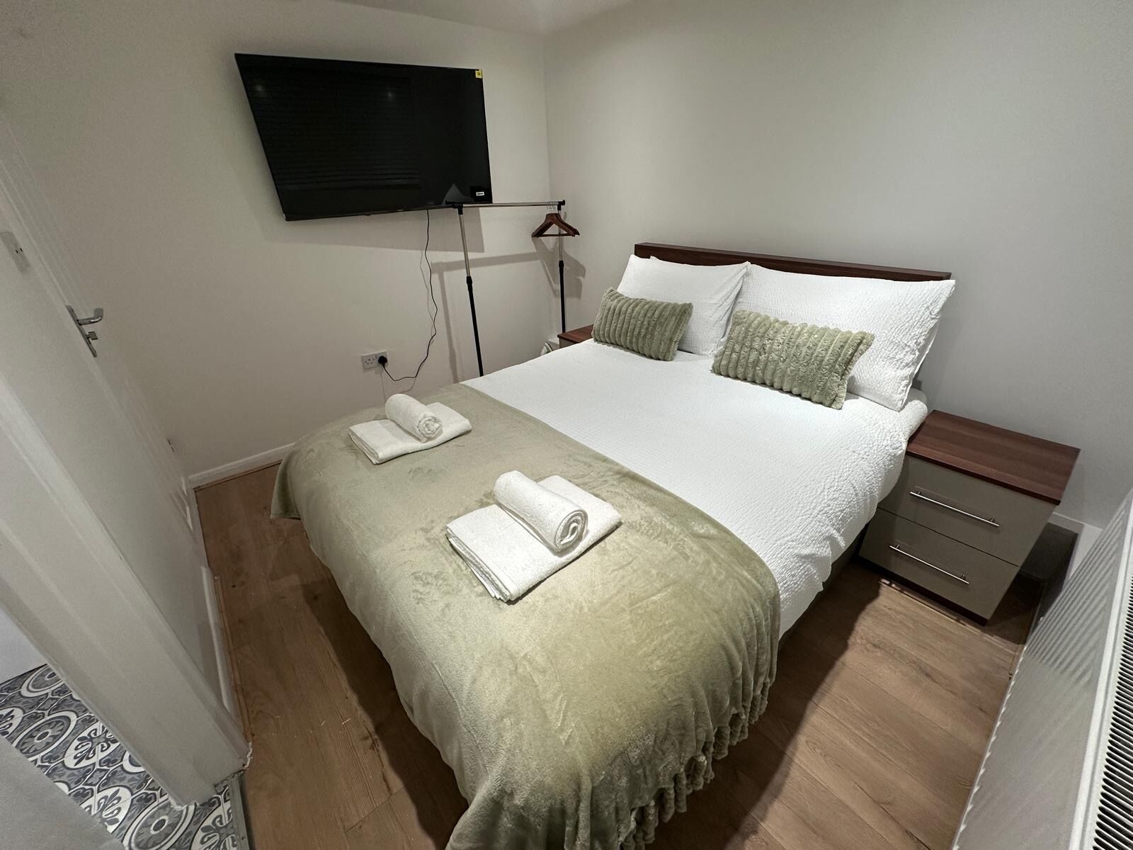 A hotel room with a bed, a TV mounted on the wall, a bedside table, and a heater.