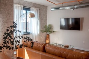 A modern living room with a leather sofa, a glass coffee table, a large flat-screen TV mounted on the wall, and a potted plant. - Commodore Barry Suite (Philadelphia)