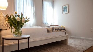 Quarto com uma cama, mesa de cabeceira com um vaso de flores e uma janela com cortinas.