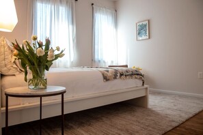 A bedroom with a bed, a bedside table with a vase of flowers, and a window with curtains. - Commodore Barry Suite (Philadelphia)
