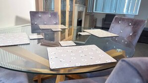 Uma mesa com tampo de vidro, jogos americanos florais, cadeiras acolchoadas e um armário de madeira ao fundo.