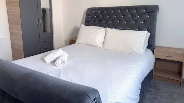 Uma cama bem arrumada com cabeceira estofada, um guarda-roupa cinzento e uma pequena mesa de cabeceira com abajur.