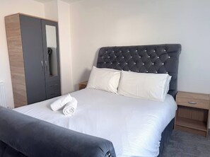 A neatly made bed with a tufted headboard, a grey wardrobe, and a small bedside table with a lamp.