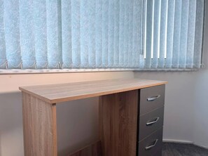 Une pièce avec un bureau en bois, un meuble à tiroirs intégré et une fenêtre équipée de stores verticaux.