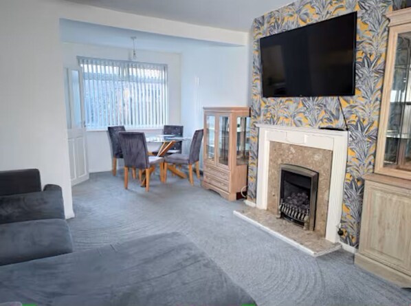 A living room with a fireplace, a television mounted on the wall, a dining table with chairs, and a sofa.