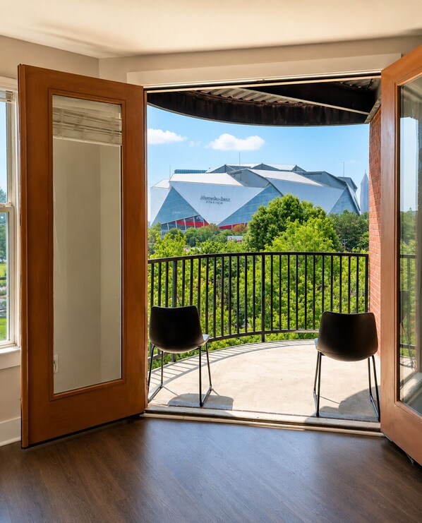 An open door leading to a balcony with two chairs, a view of a stadium, and a brick wall.