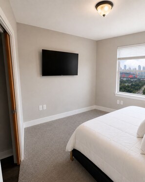 A bedroom with a bed, a flat-screen TV mounted on the wall, a window with a city view, and carpeted flooring.