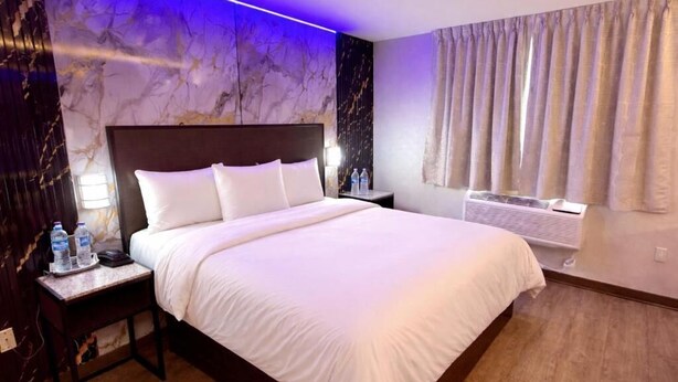A hotel room with a large bed, a nightstand with a lamp, a wall-mounted air conditioner, and a window with curtains. - Imperial Hotel (Brooklyn)