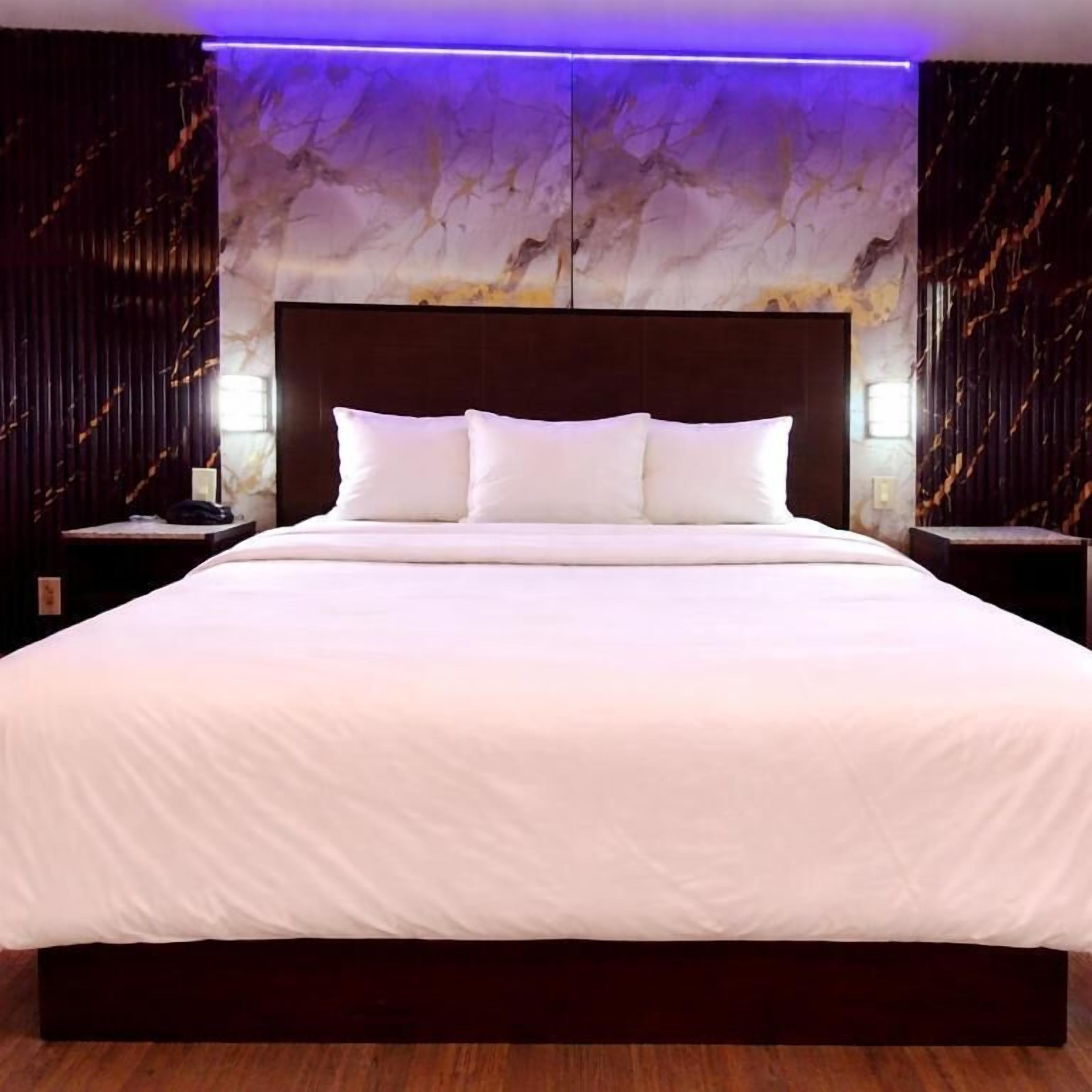 Una cama grande con ropa blanca, cabecera oscura y pared con diseño de mármol al fondo.