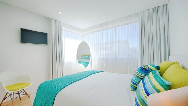 Ein modernes Hotelzimmer mit einem großen Bett, einem an der Wand montierten Fernseher, einem Hängesessel und Blick auf das Meer durch leichte Vorhänge.