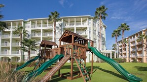 Een speeltuin met glijbanen en klimconstructies voor een hotel met meerdere balkons en palmbomen.