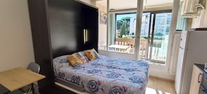 A bedroom with a bed, a small table, a refrigerator, and a balcony with a view of buildings. - Studio with Balcony, Pool & Parking. (Fréjus)