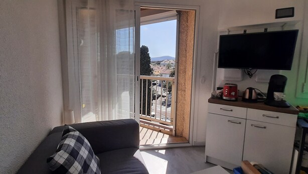 Uma varanda com vista para edifícios e uma montanha, um sofá com almofada xadrez, uma televisão fixada na parede e uma kitchenette com armários e uma máquina de café.
