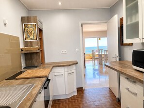 A kitchen with white cabinets, a wooden countertop, and a view of the sea through an open door. - Apartment with Panoramic Sea View (Ajaccio)