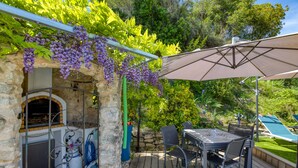 A stone-built outdoor kitchen with a grill, a table with chairs, and a pergola with hanging vines. - Les Vignes Guest Room with Private Entrance, Wi-Fi, and Air Conditioning (Puisserguier)
