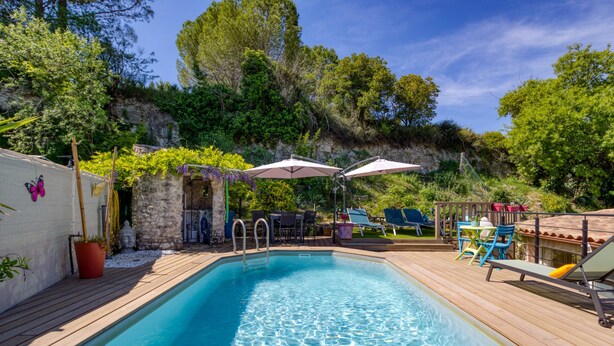 A swimming pool with a wooden deck, surrounded by greenery and a stone wall. - Les Vignes Guest Room with Private Entrance, Wi-Fi, and Air Conditioning (Puisserguier)