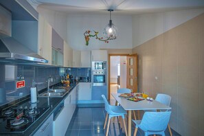A modern kitchen with a gas stove, oven, and a dining area with a table and chairs. - Savana Desert View Apartment – ​​Sea View Super King Bed Private Parking Sleeps 7  (Caniço)