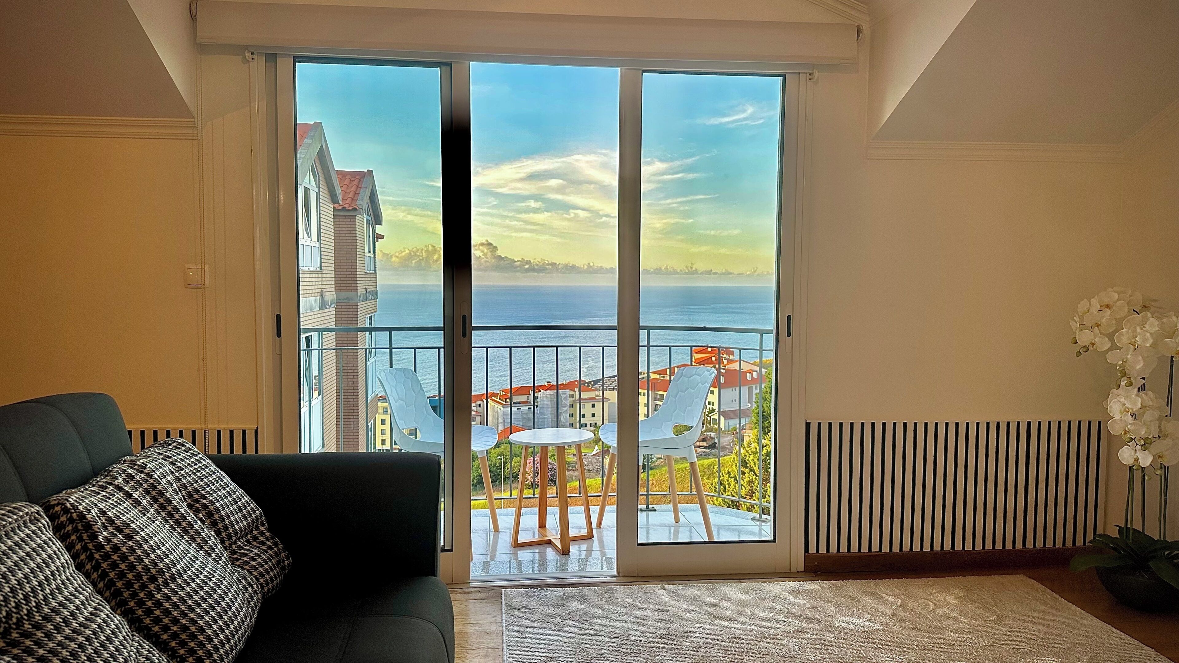 A living room with a sofa, a view of the sea through a sliding glass door, and a potted plant.
