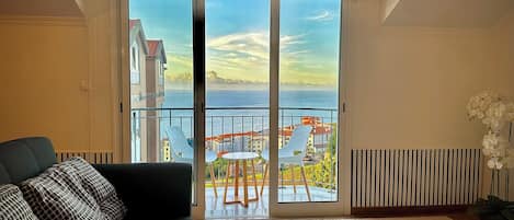 A living room with a sofa, a view of the sea through a sliding glass door, and a potted plant.