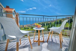 Un balcone con due sedie bianche, un tavolino rotondo con una ciotola di frutta e vista sul mare e sugli edifici.