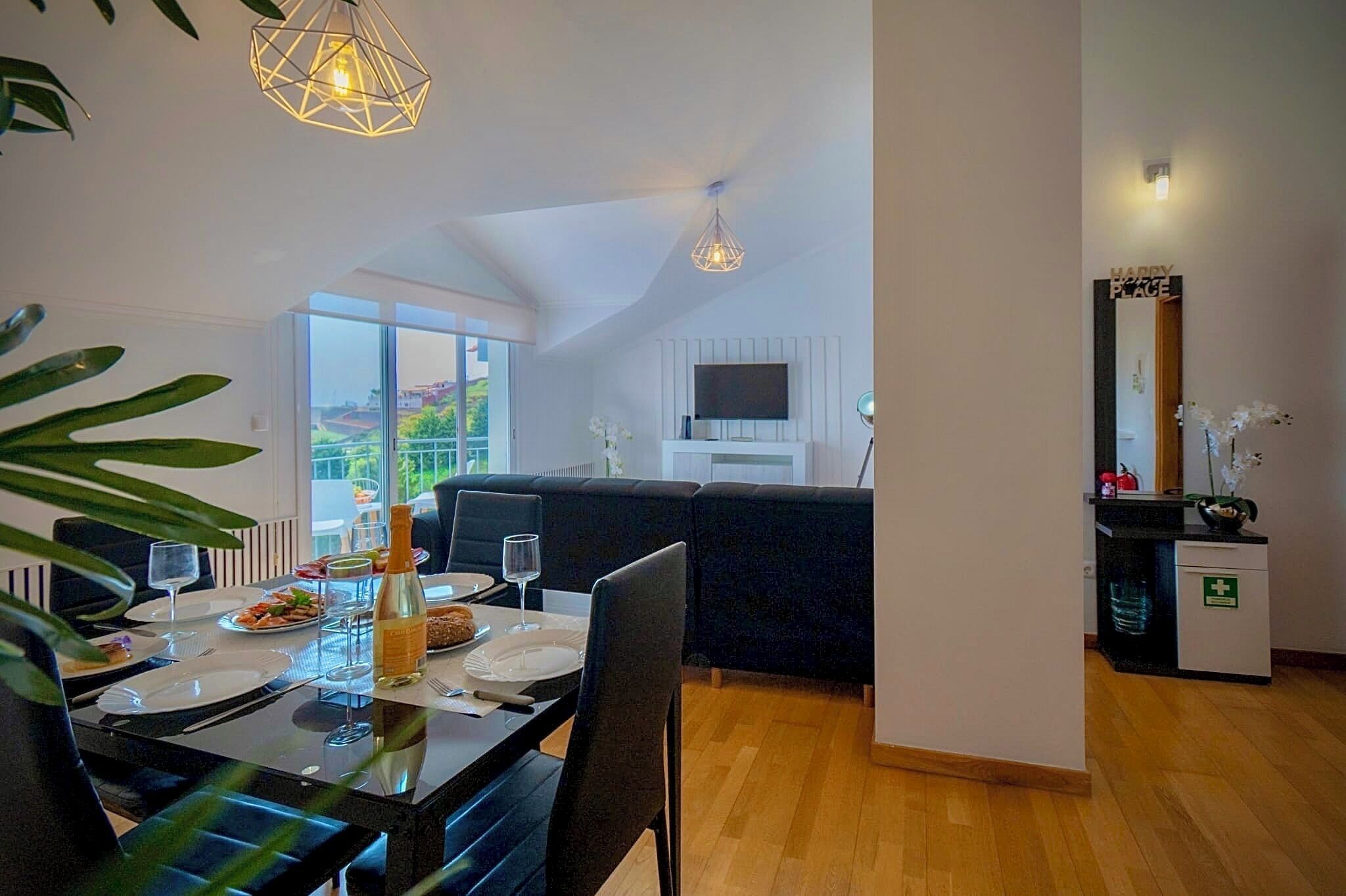 A dining area with a table set for two, a sofa, a TV, and a small kitchenette with a mirror and a sign.