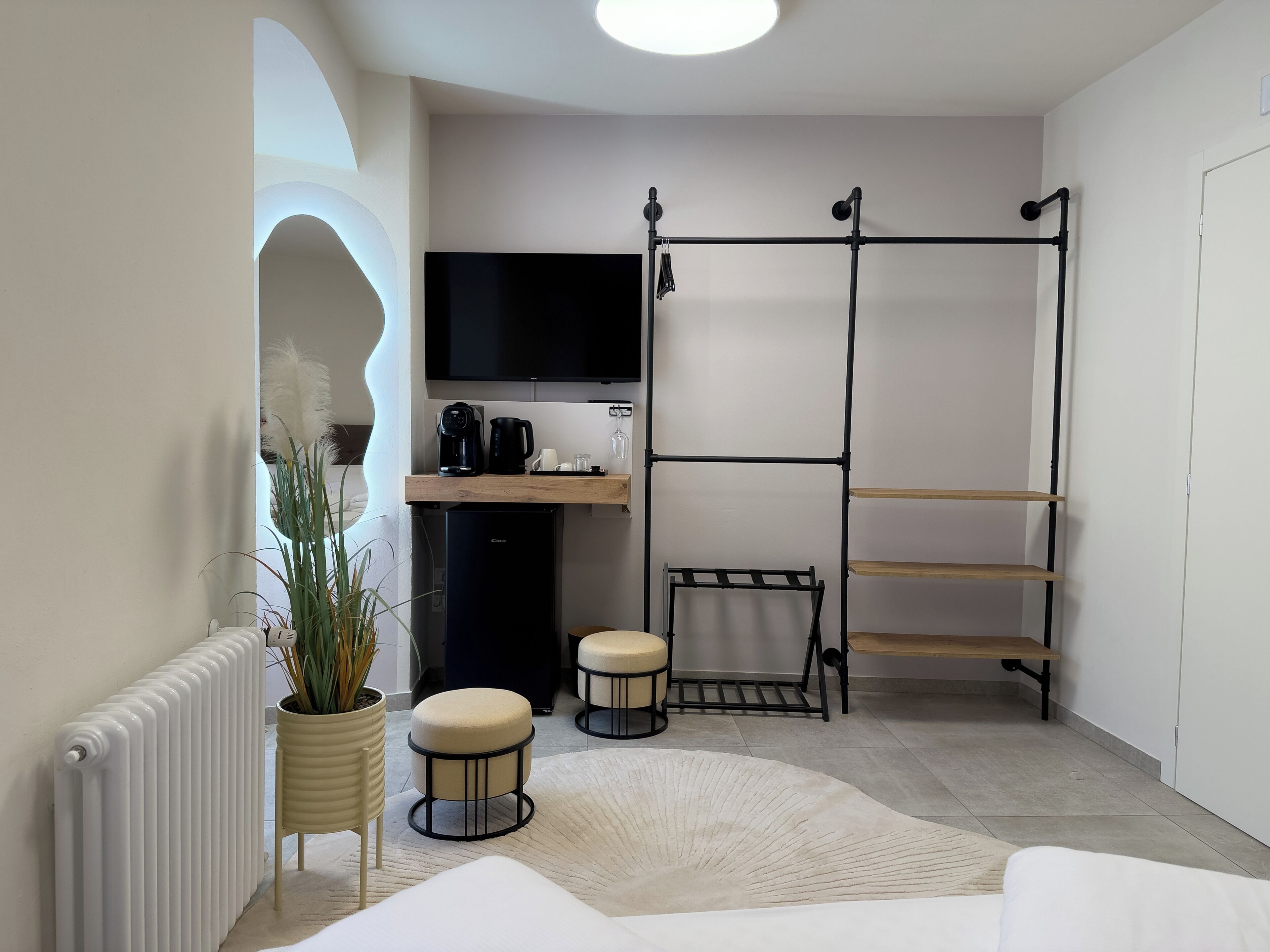 Ein kleines Zimmer mit Küchenzeile, einem an der Wand befestigten schwarzen Fernseher, einer Heizung, einer Topfpflanze, zwei Hocker und einem weißen Sofa.