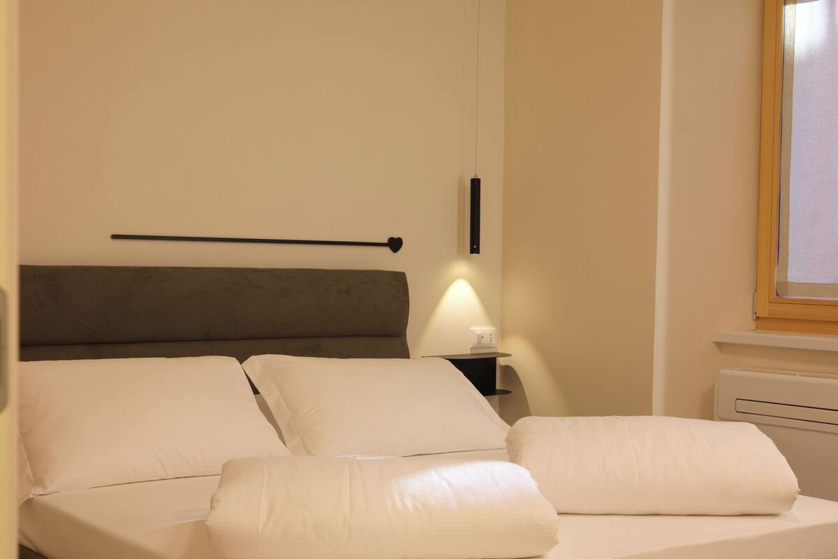 A neatly made bed with white linens, a bedside table with a lamp, and a wall-mounted light fixture.