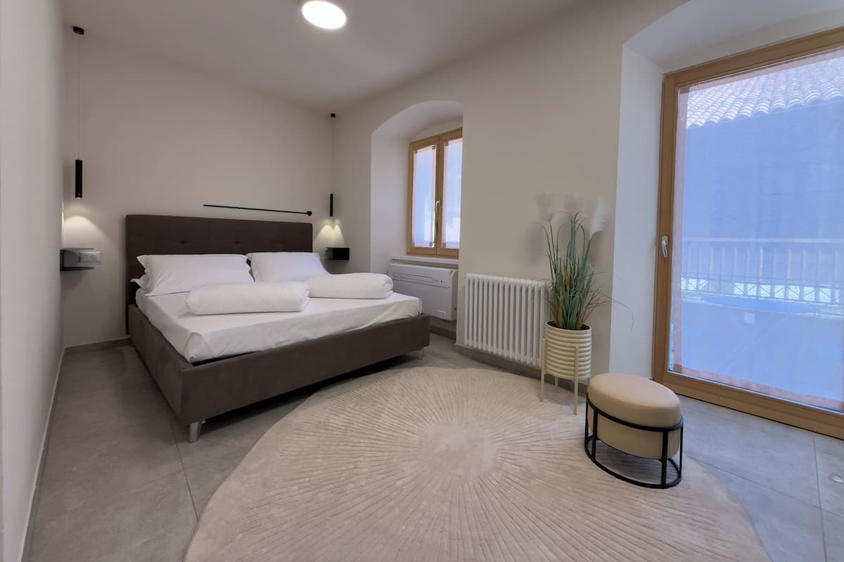 A bedroom with a bed, bedside lamps, a radiator, a window, and a view of a balcony.