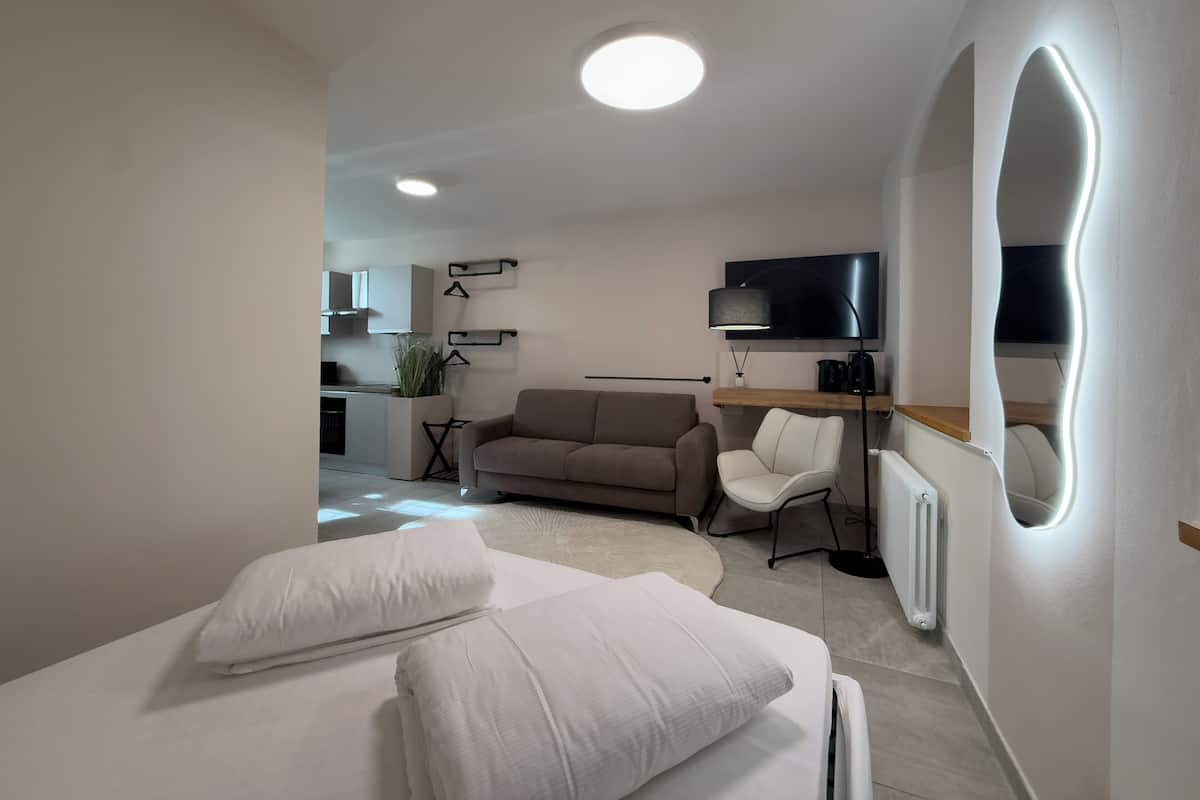 A modern hotel room with a bed, a sofa, a desk with a computer, a TV, and a kitchen area in the background.