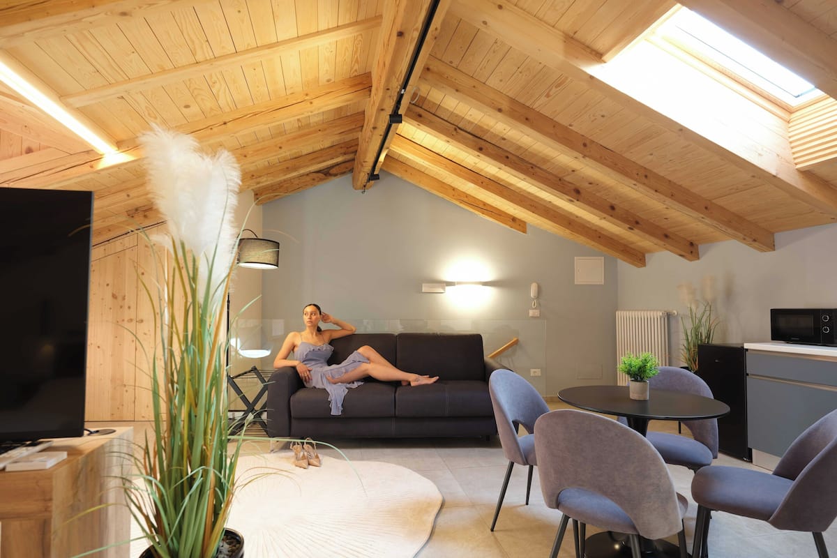 A modern loft with a wooden ceiling, a sofa, a dining table, chairs, a TV, and a kitchen area.