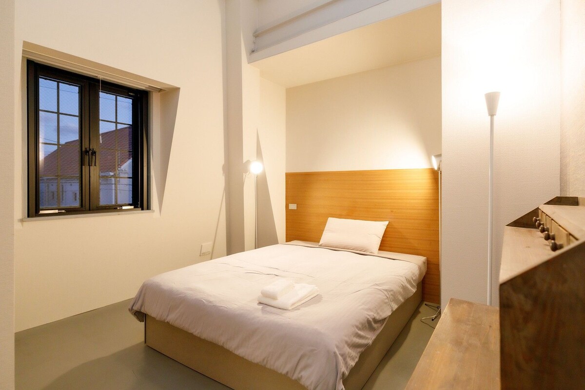 A hotel room with a bed, a window, a headboard, a bedside table, and a staircase.