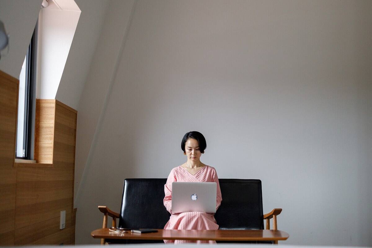 A person seated on a black chair with a laptop, in a room with a sloped ceiling and wooden paneling.