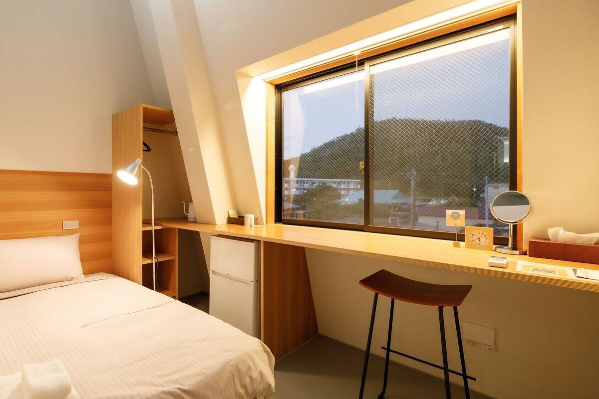 A bedroom with a bed, a desk, a chair, a window, and a small kitchenette.