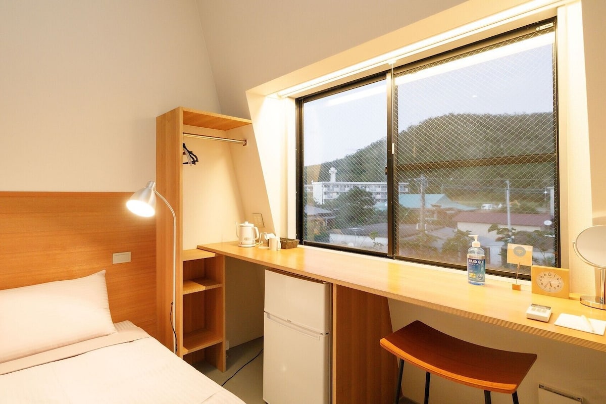 A compact room with a bed, a desk, a chair, a window with a view, and a small shelf.